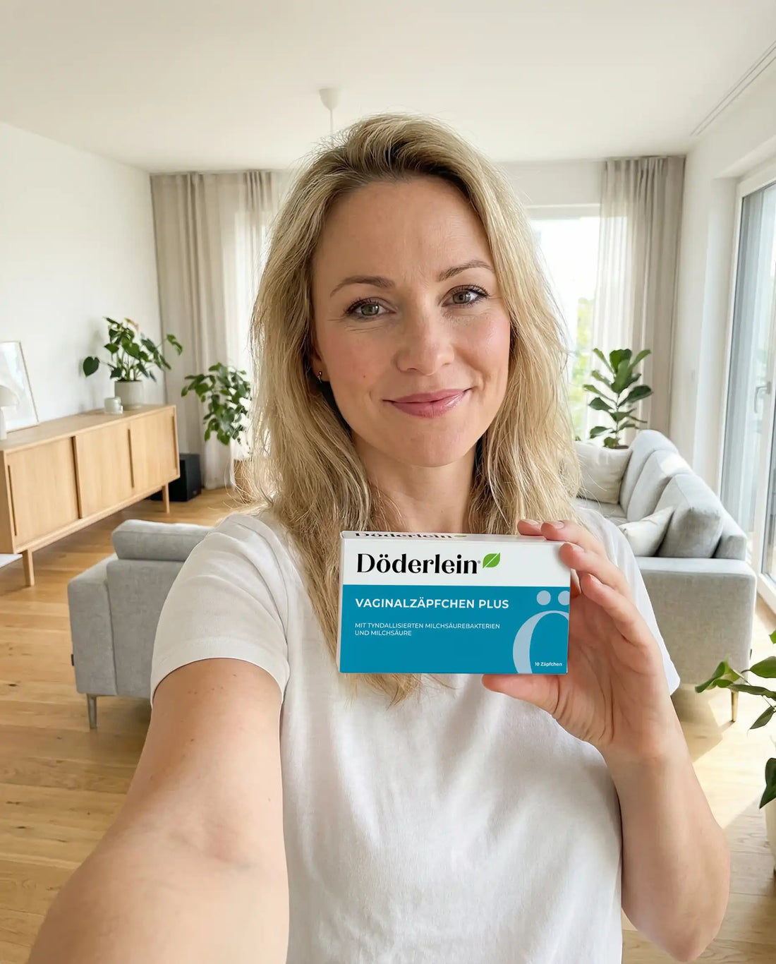 Eine lächelnde Frau mit blondem Haar steht in einem hellen Wohnzimmer und hält eine Schachtel mit der Aufschrift Döderlein Vaginalzäpfchen Plus in die Kamera. Sie trägt ein weißes Hemd, und im Hintergrund sind Pflanzen und Möbel zu sehen.